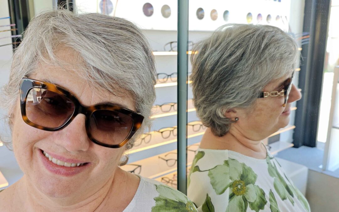 Una donna,Ludovica, sorridente con capelli corti prova degli occhiali da sole con lenti graduate tartarugati con lenti sfumate all'interno di un negozio di ottica. Alla sua destra, uno specchio riflette il suo profilo, mostrando le aste degli occhiali e una maglietta a motivi floreali verdi e bianchi. Sullo sfondo si vedono espositori illuminati con varie montature da vista.