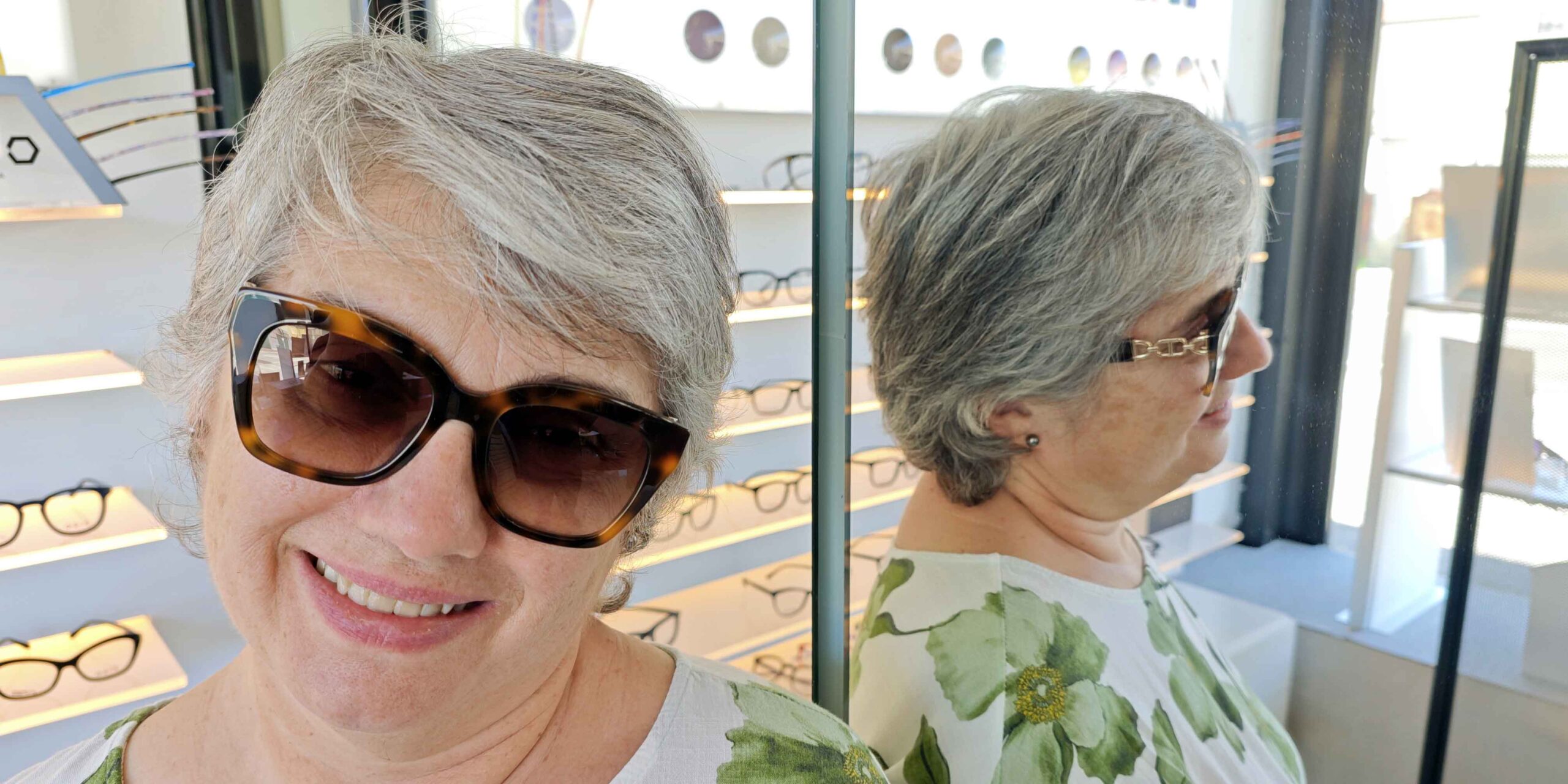 Una donna,Ludovica, sorridente con capelli corti prova degli occhiali da sole con lenti graduate tartarugati con lenti sfumate all'interno di un negozio di ottica. Alla sua destra, uno specchio riflette il suo profilo, mostrando le aste degli occhiali e una maglietta a motivi floreali verdi e bianchi. Sullo sfondo si vedono espositori illuminati con varie montature da vista.