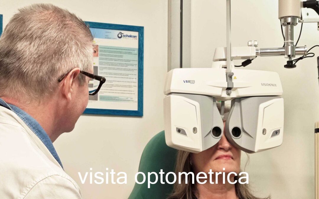Una cliente è seduta su una sedia medica verde mentre guarda attraverso un grande forottero digitale bianco di marca Visionix per la misurazione della vista. Sulla sinistra, un ottico di spalle con occhiali e camice bianco monitora la procedura. Sullo sfondo si notano due cartelli informativi incorniciati con il logo Schalcon appesi alla parete bianca. In basso al centro è presente la scritta in sovrimpressione bianca "visita optometrica".
