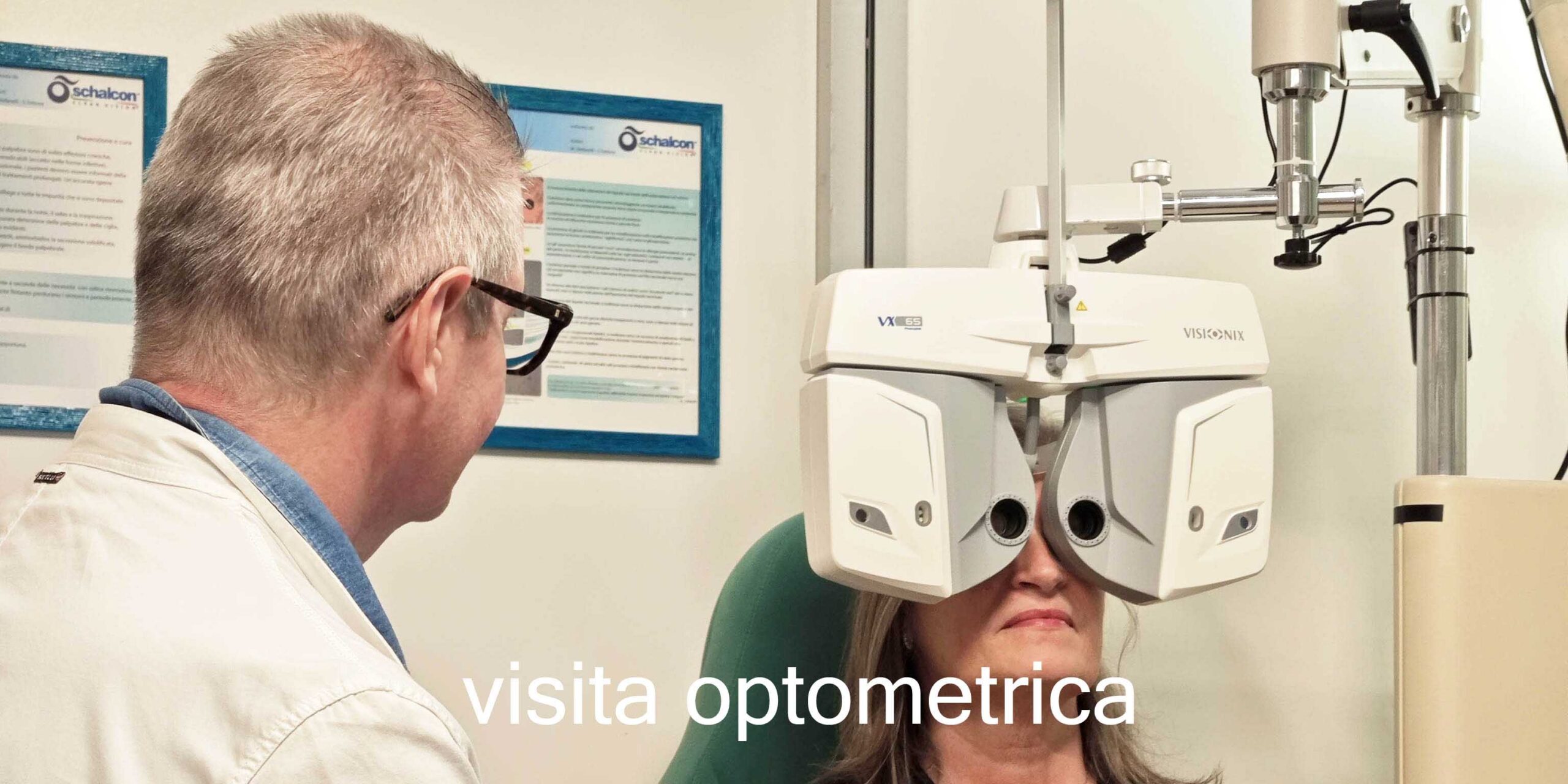 Una cliente è seduta su una sedia medica verde mentre guarda attraverso un grande forottero digitale bianco di marca Visionix per la misurazione della vista. Sulla sinistra, un ottico di spalle con occhiali e camice bianco monitora la procedura. Sullo sfondo si notano due cartelli informativi incorniciati con il logo Schalcon appesi alla parete bianca. In basso al centro è presente la scritta in sovrimpressione bianca "visita optometrica".